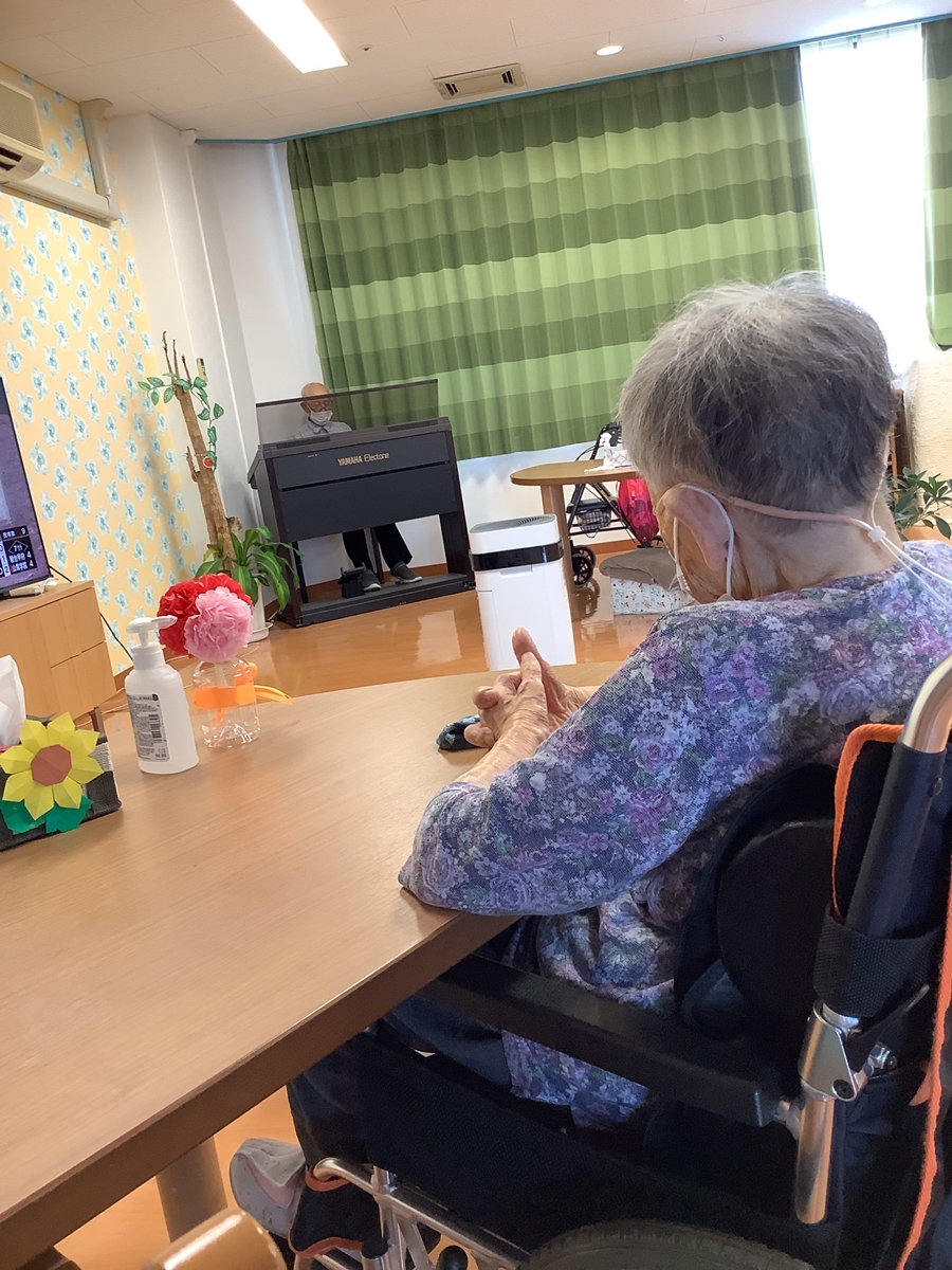 高齢者が室内でピアノを楽しむレクリエーション風景。電子ピアノと花の装飾がある活動スペース。